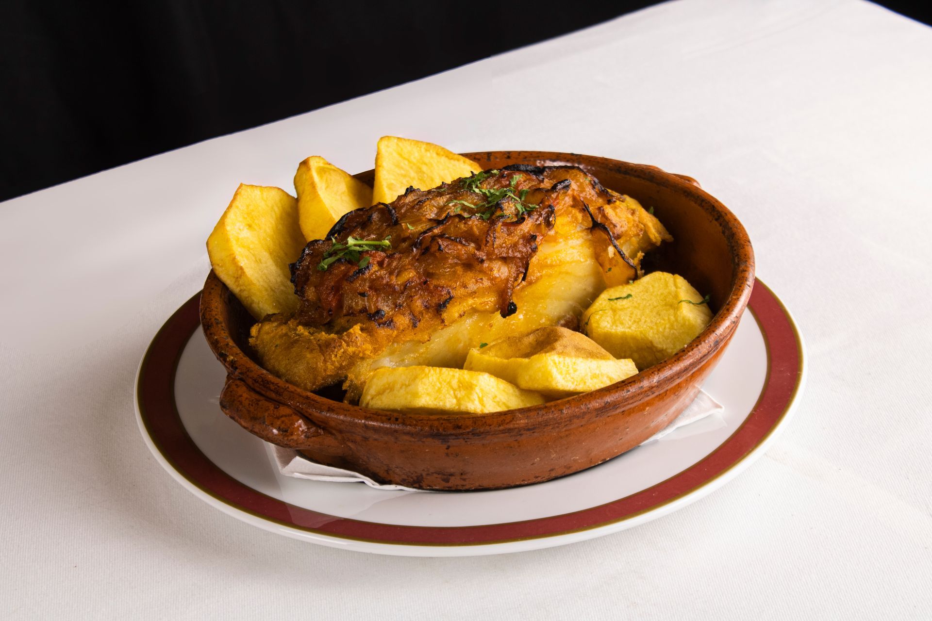 Peixe assado e batatas em um prato marrom, sobre uma toalha de mesa branca.