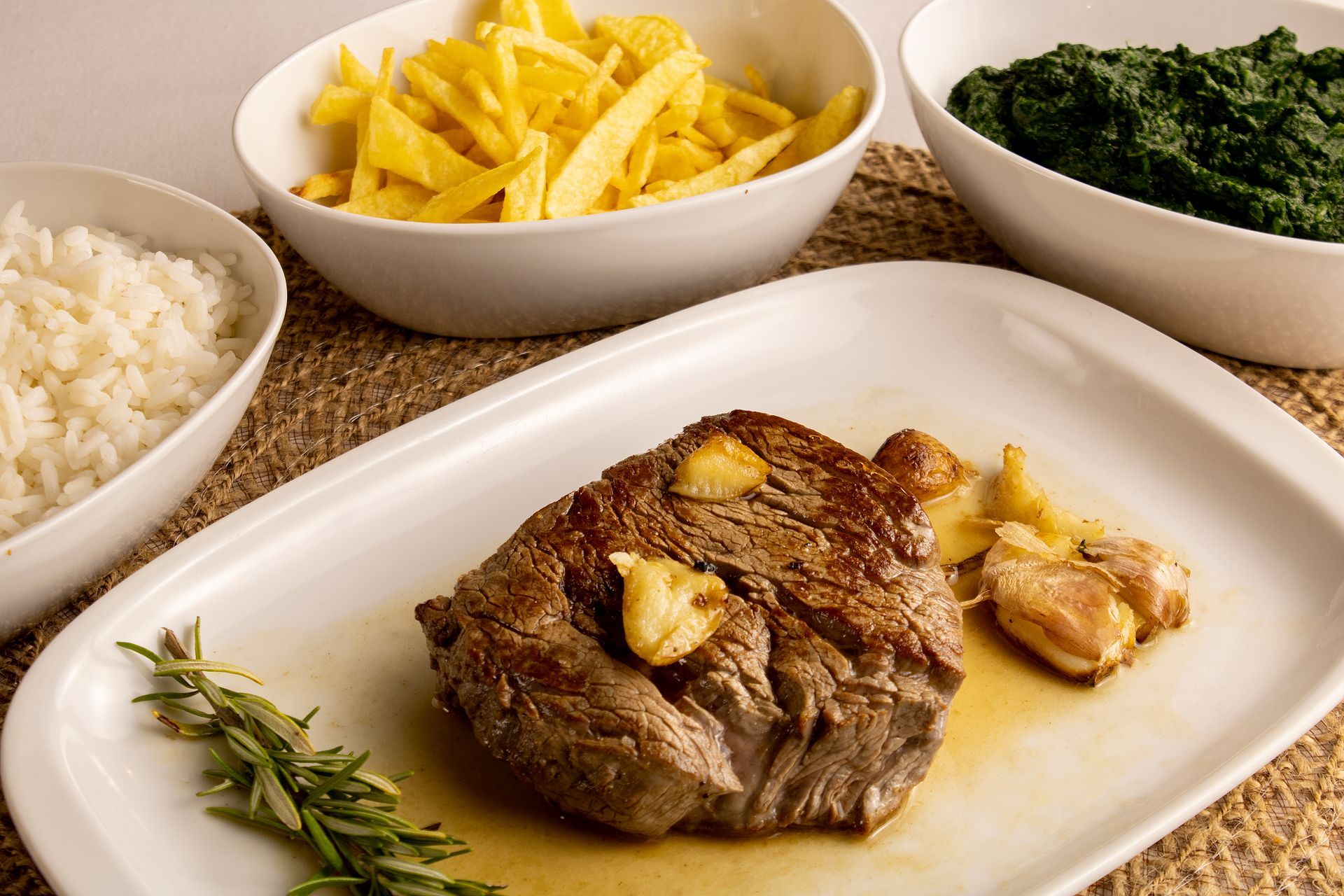 Bife com alho em um prato branco, servido com arroz, batatas fritas e espinafre em tigelas.