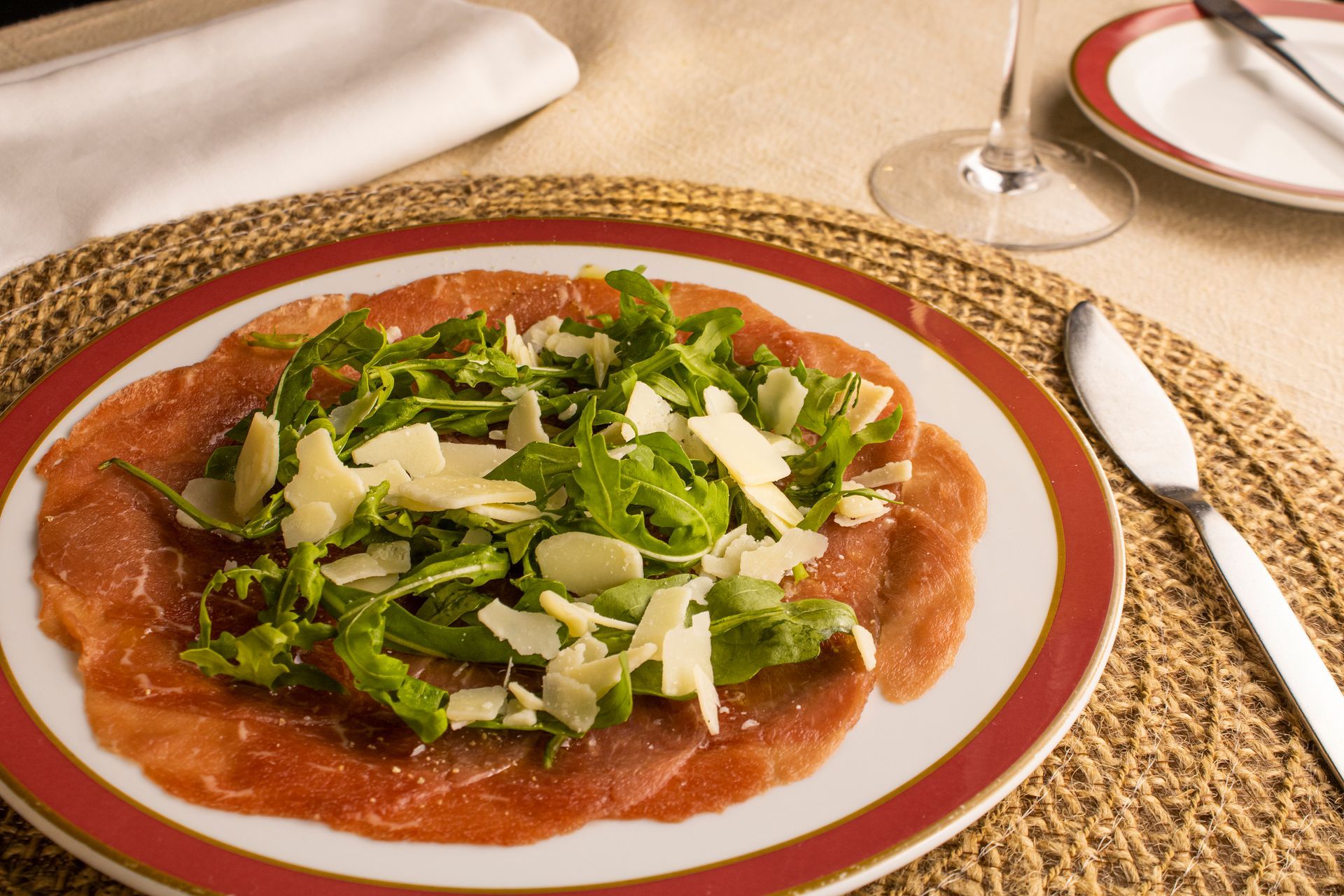 Carpaccio com rúcula e queijo parmesão em um prato com borda vermelha, servido à mesa.