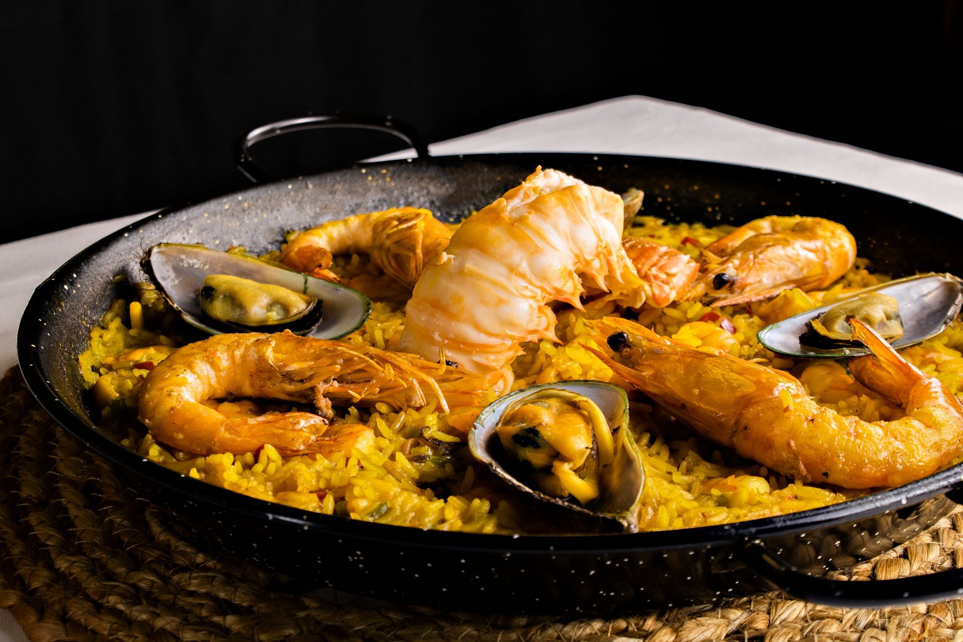 Paella com camarão, mexilhões e cauda de lagosta em uma panela preta sobre uma esteira de tecido.