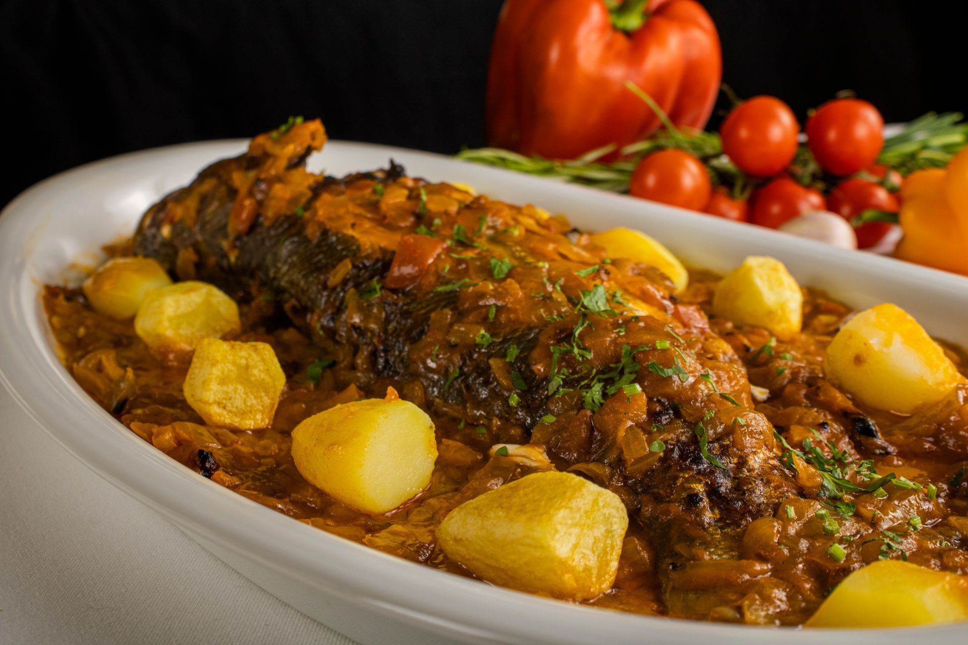 Peixe inteiro cozido com batatas em um prato oval branco, guarnecido com ervas frescas e tomates.
