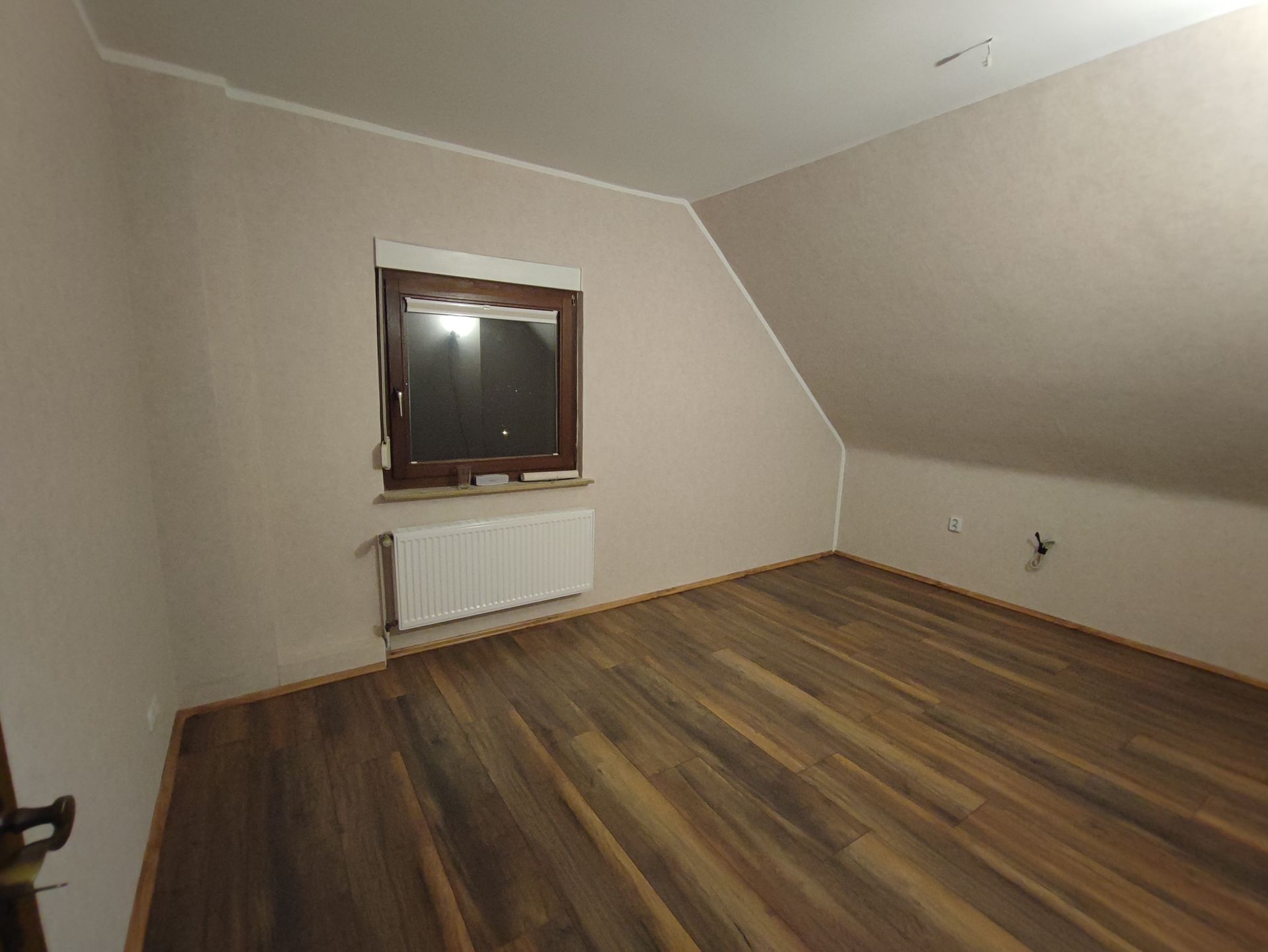 Leeres Zimmer mit Holzboden, hellbraunen Wänden, Dachschräge, Fenster und Heizkörper.