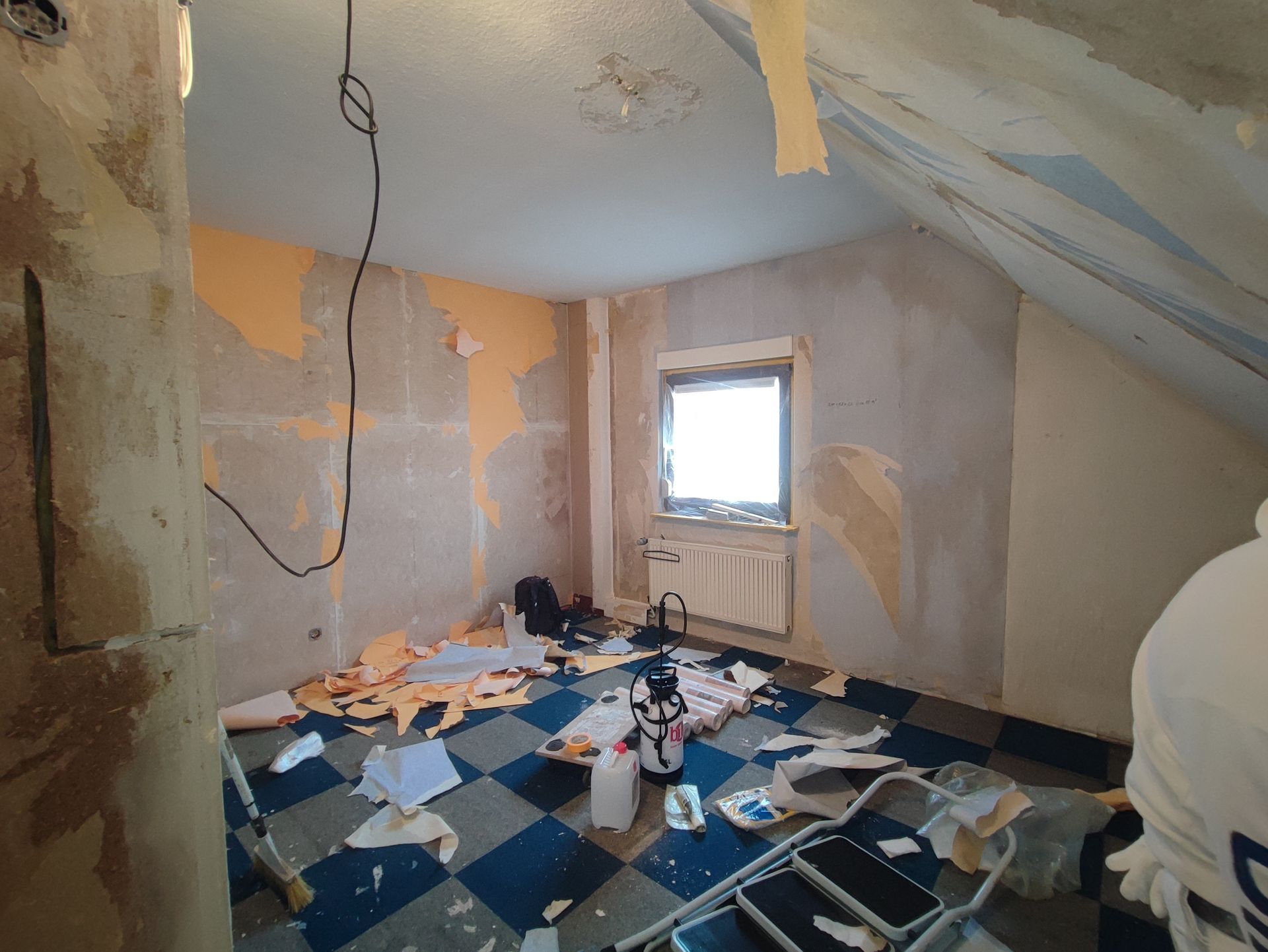 Zimmer wird renoviert: Wände mit entfernter Tapete, Schutt auf dem blau-weiß karierten Boden, kleines Fenster.