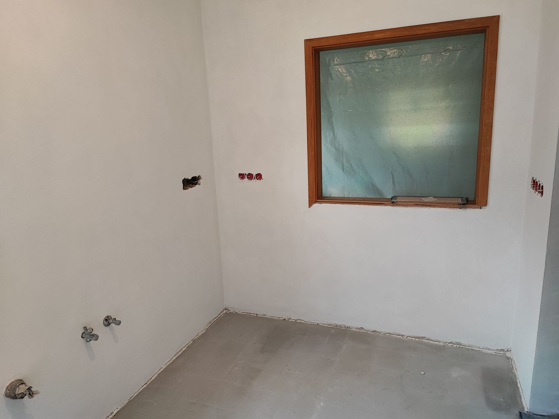 Innenecke eines Zimmers mit weißen Wänden, Betonboden, Fenster und Steckdosen.