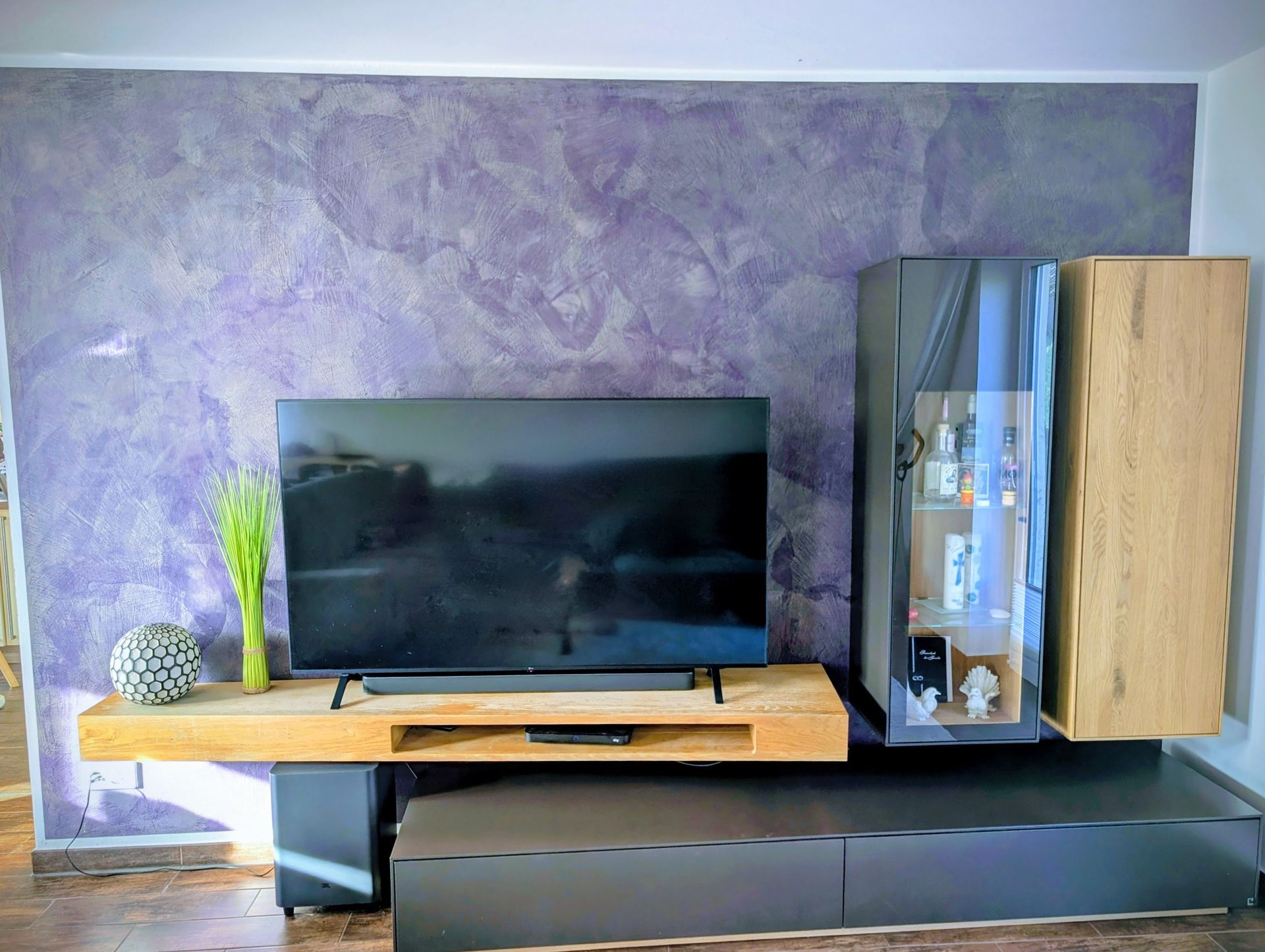 Fernseher und schwebendes Holzregal vor einer strukturierten, violetten Wand, dazu dunkelgraue und holzfarbene Schränke.