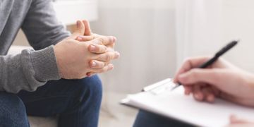 Un hombre está sentado en un sofá hablando con una mujer que está escribiendo en un portapapeles.