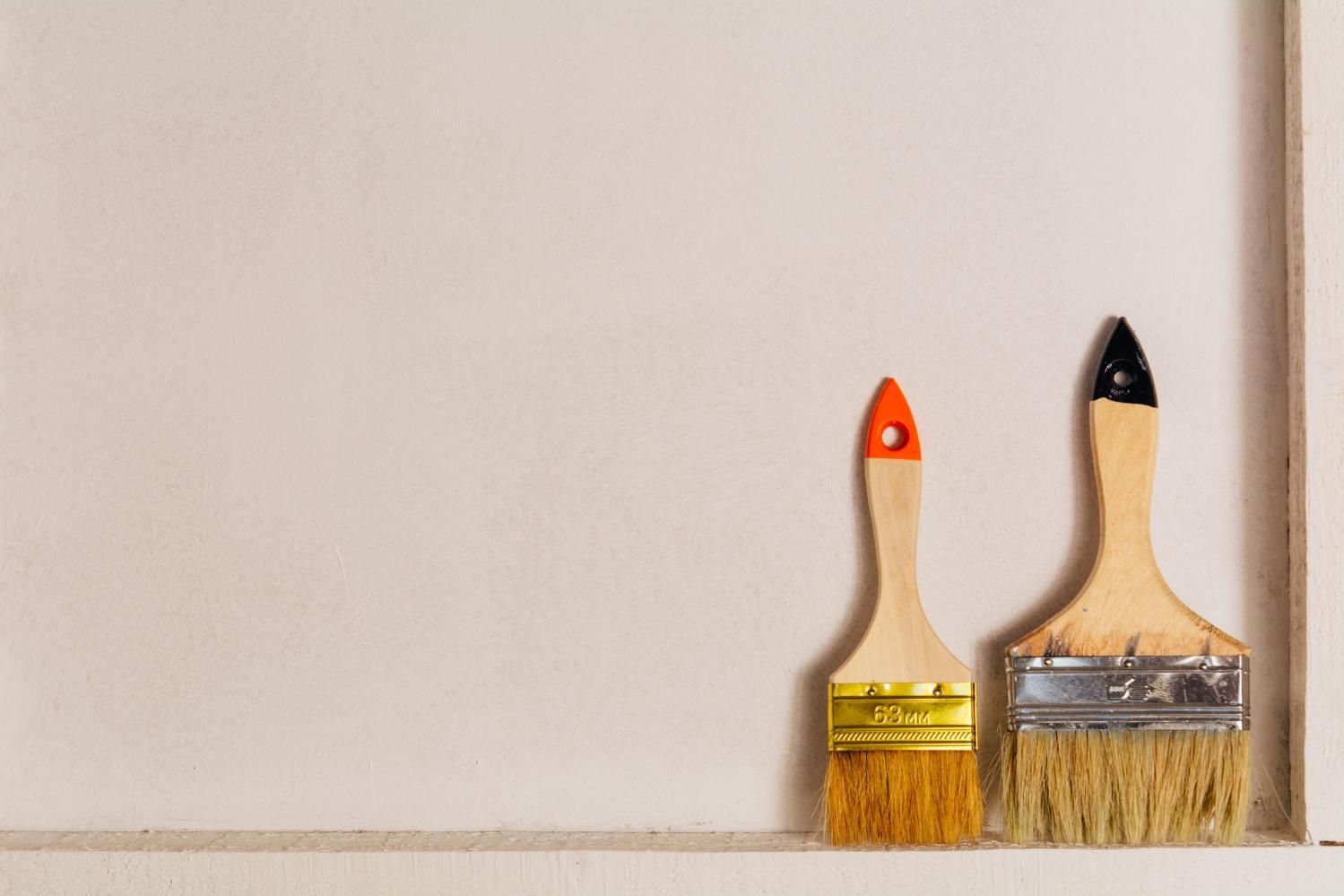 Dos pinceles apoyados contra una pared, uno pequeño con cerdas naranjas y otro más grande con cerdas color canela.