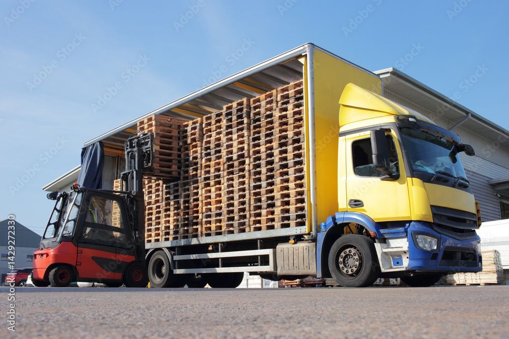 Rote und gelbe Gabelstapler heben gestapelte Holzpaletten neben einem blau-gelben Lkw auf einem Lagerplatz an.