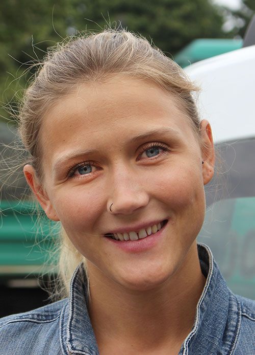 Blonde Frau lächelt, trägt eine Jeansjacke, einen Nasenring und ihr Haar ist zurückgekämmt. Im Freien, teilweise sichtbarer Hintergrund.
