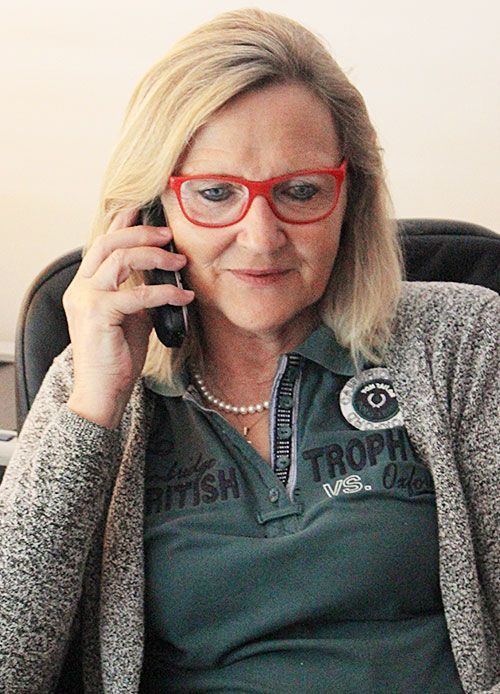 Frau mit blonden Haaren und roter Brille am Telefon. Trägt grünes Poloshirt, Perlenkette und graue Strickjacke.