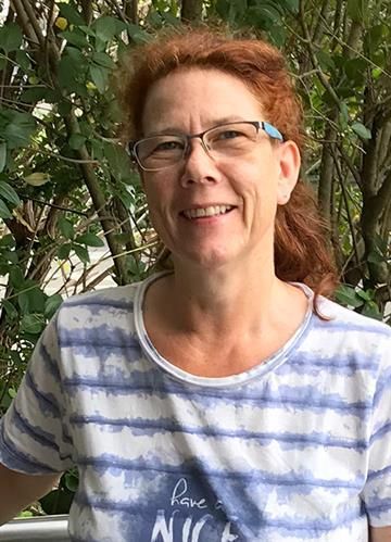 Frau mit roten Haaren und Brille lächelt im Freien in die Kamera und trägt ein gestreiftes blau-weißes Hemd.
