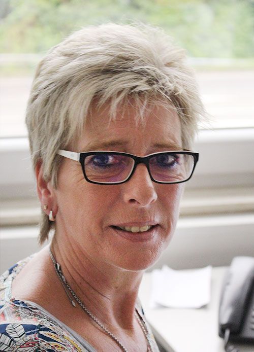 Frau mit kurzen blonden Haaren, Brille, lächelnd, in einem Büro.