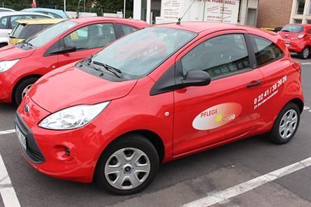Auf einem Parkplatz steht ein roter Ford Ka mit einem Logo an der Seite, dahinter steht ein weiteres rotes Auto.