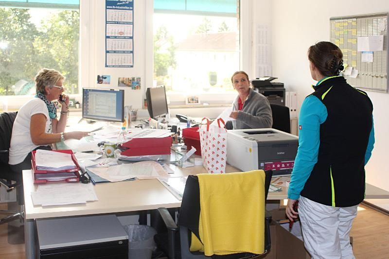 Drei Personen in einem Büro; Frau am Telefon, Mann am Schreibtisch, Person daneben, helle Büroumgebung.