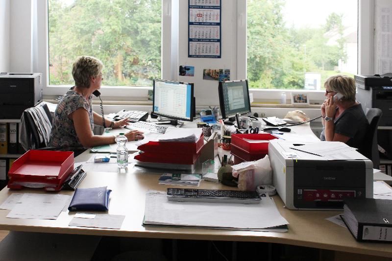 Zwei Frauen sitzen an Schreibtischen in einem Büro, telefonieren und schauen auf Computerbildschirme. Auf den Schreibtischen liegen Papiere, Drucker und Ordner.