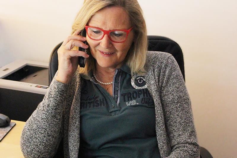 Frau mit roter Brille am Telefon, graue Strickjacke und grünes Hemd, in einem Büro.