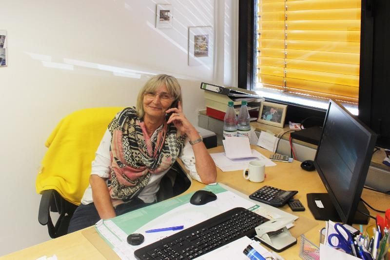Frau am Telefon am Schreibtisch, blondes Haar, trägt ein Kopftuch. Büroumgebung, leuchtend gelbe Jalousien und ein Computermonitor.