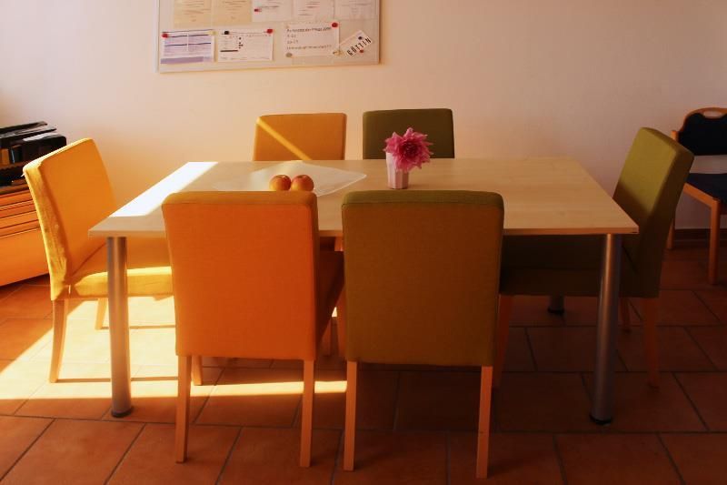 Esstisch mit sechs Stühlen, einige orange und einige grün. Auf dem Tisch stehen Blumen und Obst.