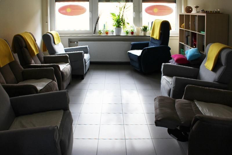 Wartezimmer mit Sesseln, gelben Decken und einem Fenster mit Pflanzen.