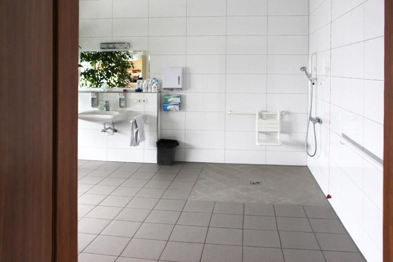 Barrierefreie Toilette mit Dusche, Waschbecken und Handläufen. Weiße Fliesen und grauer Bodenbelag.
