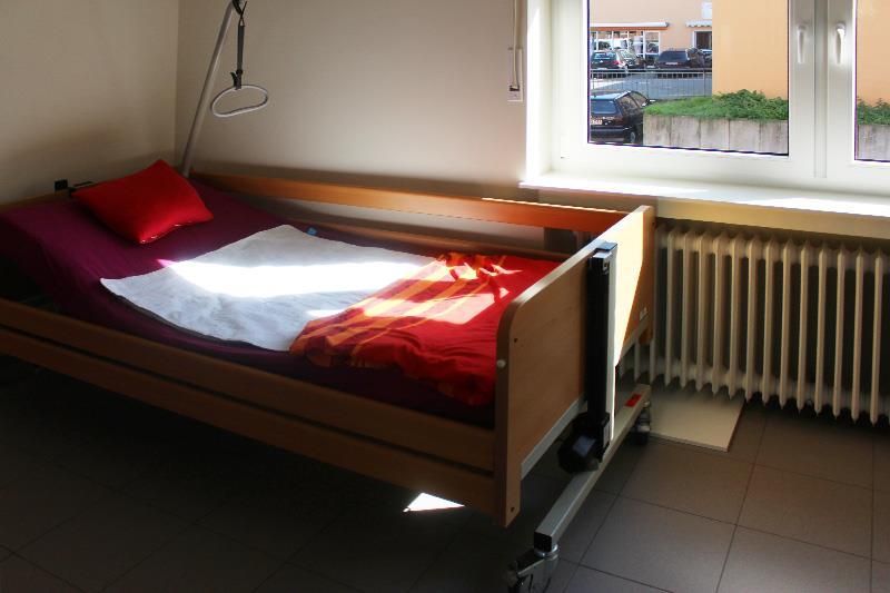 Krankenhausbett mit einem roten Kissen, einem weißen Laken und einer roten Decke in einem Zimmer mit Fenster und Heizkörper.