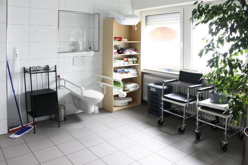 Barrierefreies Badezimmer mit Toilette, Regalen, zwei Rollstühlen und Pflanze.