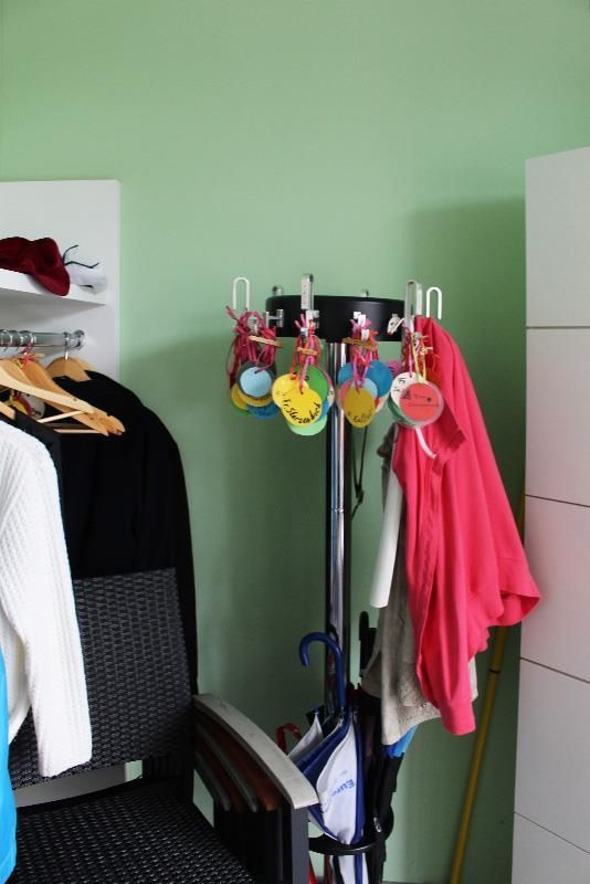 Eine Garderobe mit Kleidung und bunten Hängegegenständen, neben einem Stuhl und einer Wand.