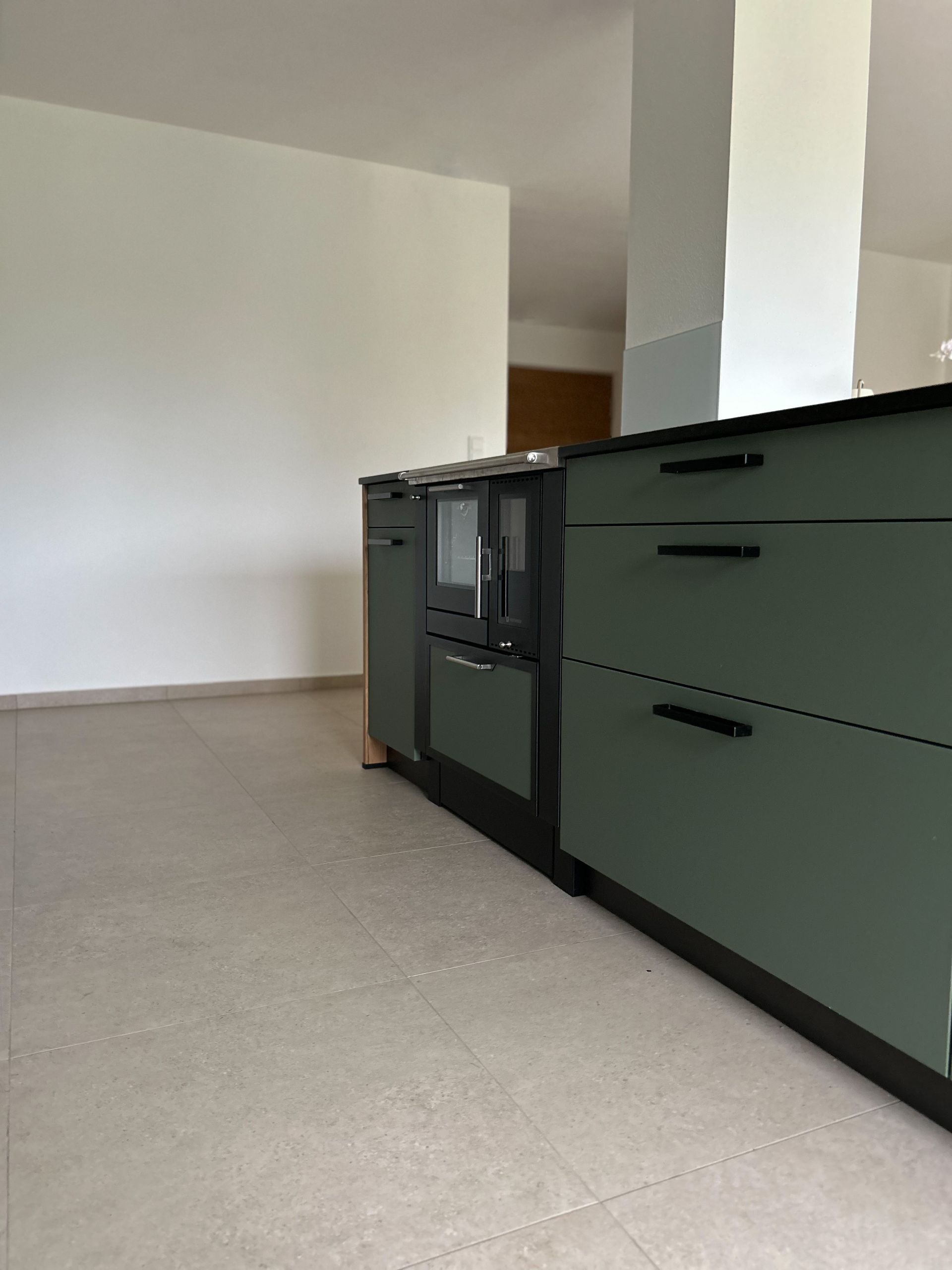 Grüne Küchenschränke mit schwarzen Akzenten. Ein kleiner Backofen ist zu sehen. Grauer Boden und weiße Wände.