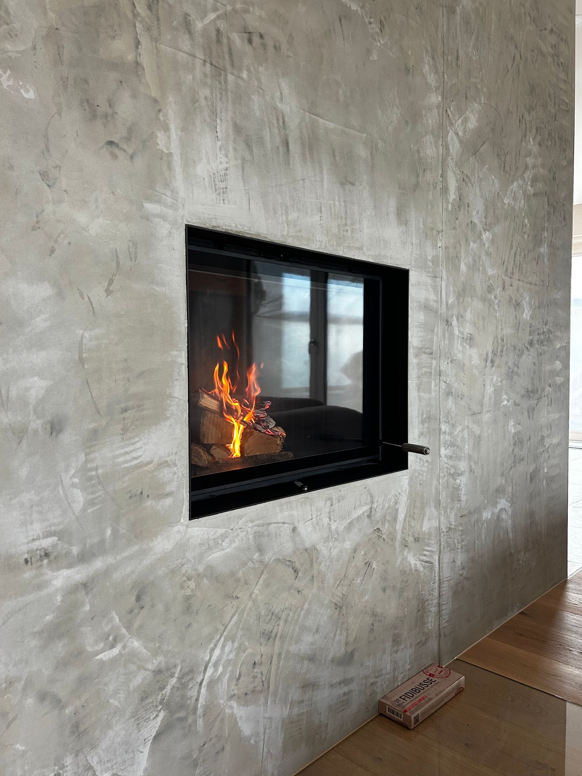 Kamin mit brennenden Flammen in einer grauen Betonwand, schwarzem Rahmen und Holzboden.