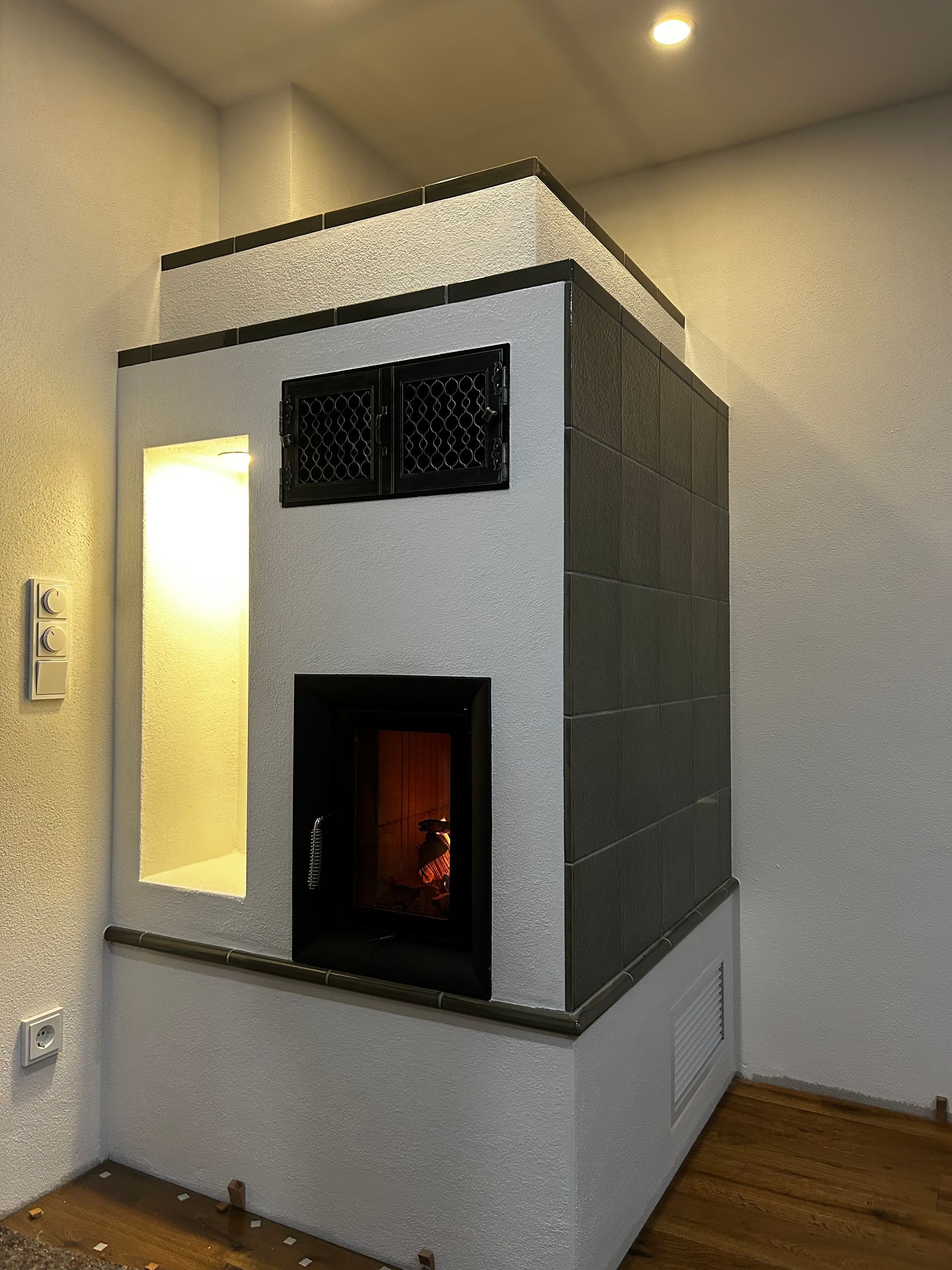 Ein weiß und dunkelgrau gefliester Kamin in einem Zimmer. Im Kamin brennt Feuer, die innere Nische ist beleuchtet.