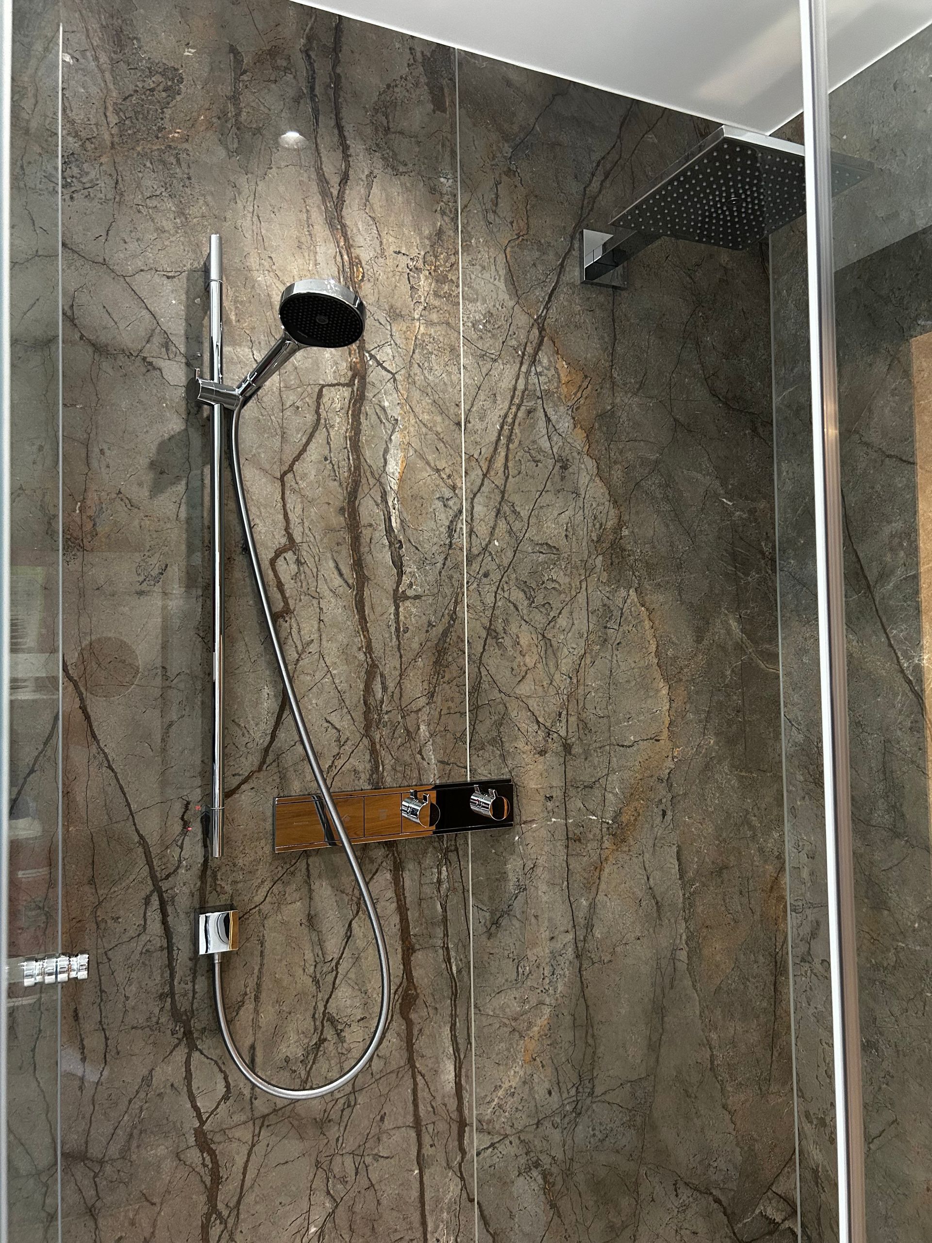 Dusche mit dunklen, marmorähnlichen Wandfliesen, zwei Duschköpfen und Glastür.