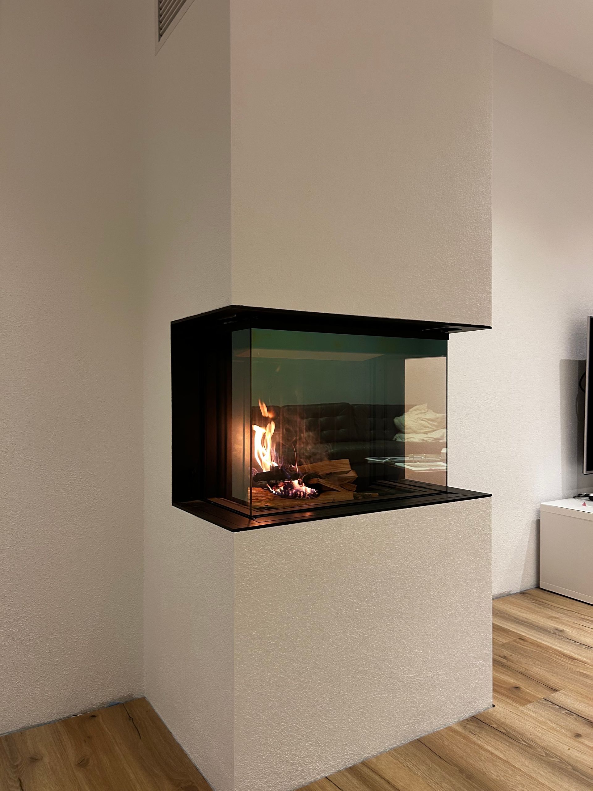 Eckkamin mit lodernden Flammen, umgeben von strukturierten weißen Wänden, Glaspaneelen und Holzböden.