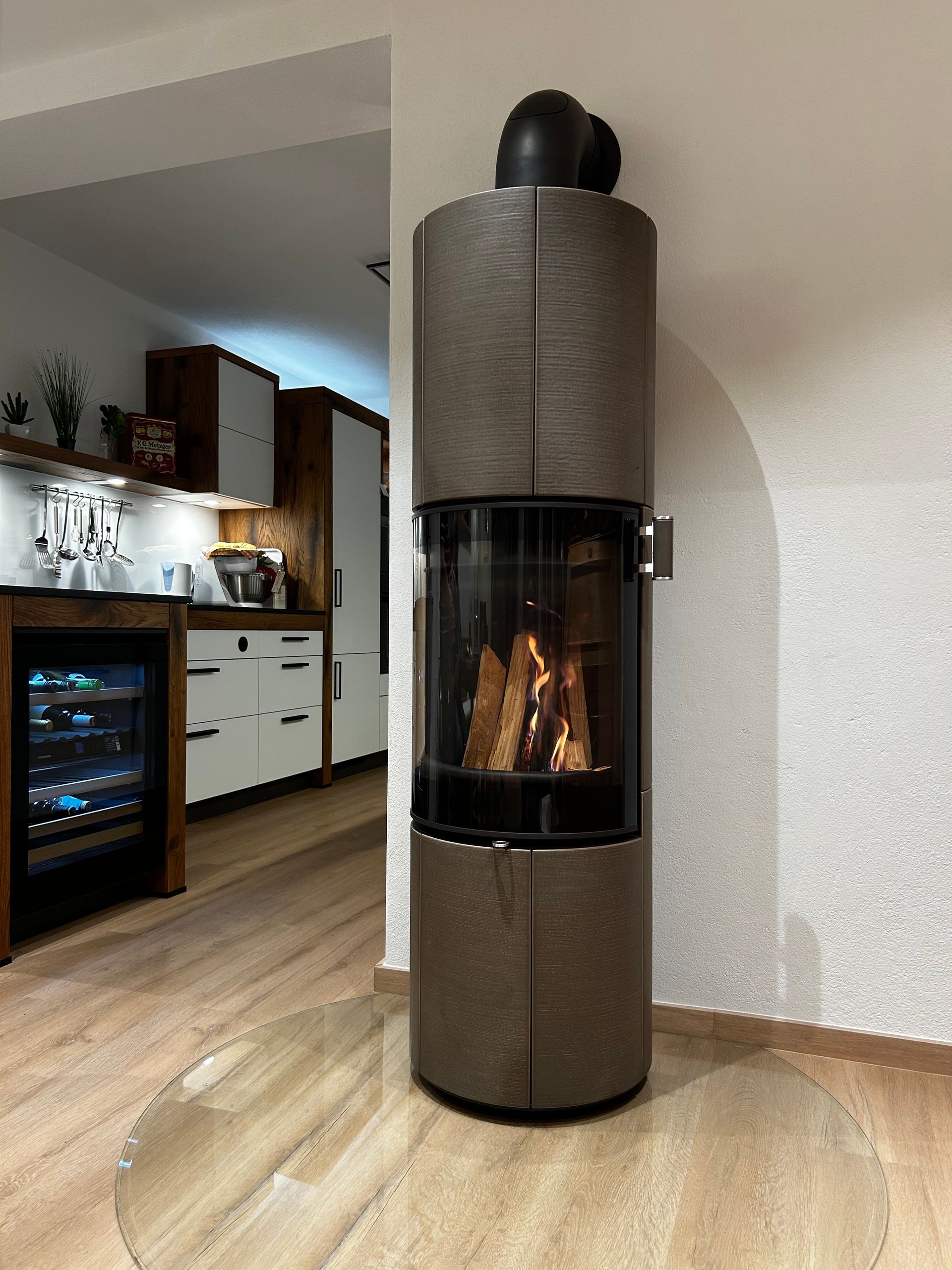 Moderner Kamin mit schwarzem Einsatz, Flammen sichtbar, eingelassen in eine weiße, strukturierte Wand, in einem lichtdurchfluteten Raum.