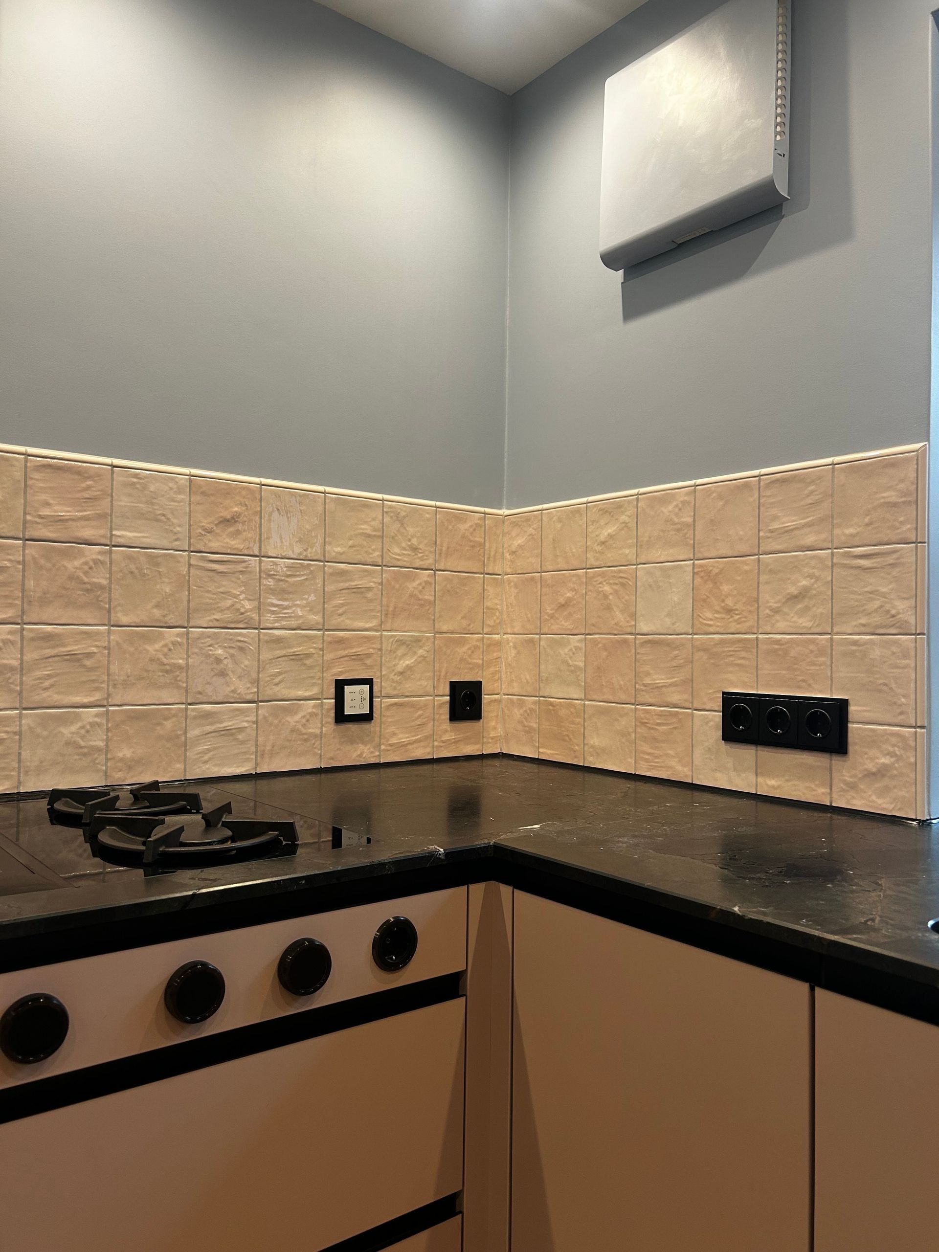 Küchenecke mit Herd, Fliesenspiegel, dunkler Arbeitsplatte und cremefarbenen Schränken. Die Wand ist grau.