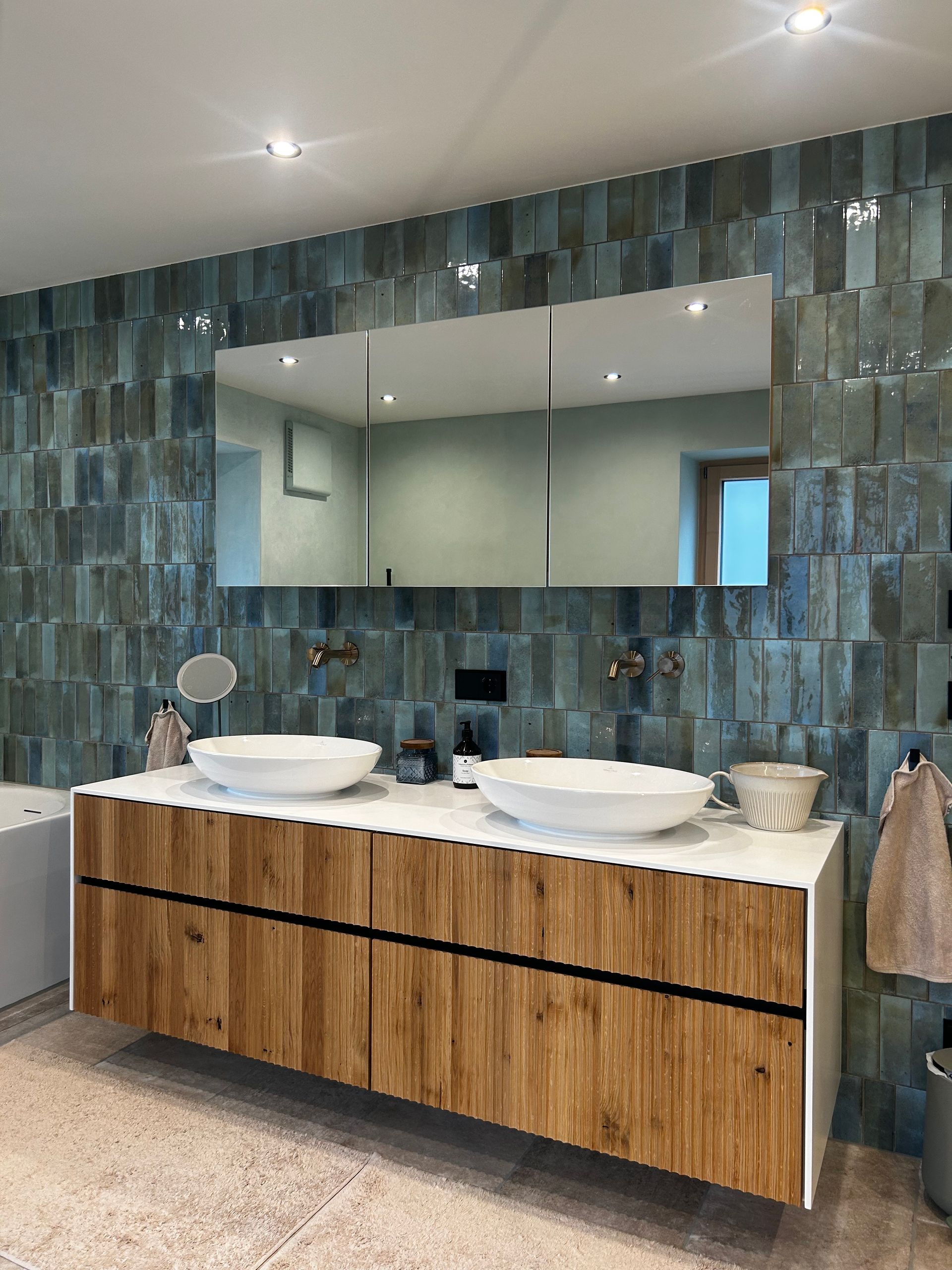 Badezimmer mit Waschtisch aus Holz, zwei Waschbecken, großem Spiegel und türkis gefliester Wand.