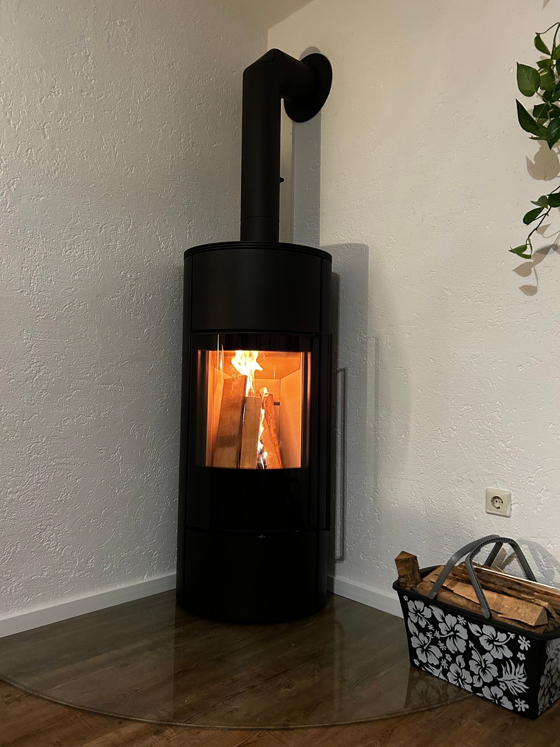 Schwarzer Holzofen mit brennendem Feuer, in einer Ecke vor einer gemusterten Wand und einem Korb.