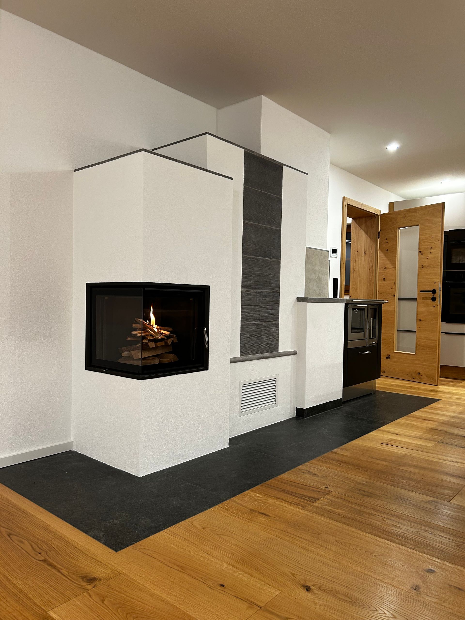 Weißer Kamin mit schwarzem Zierrand und Feuer, daneben dunkelgrauer Stein- und Holzboden.