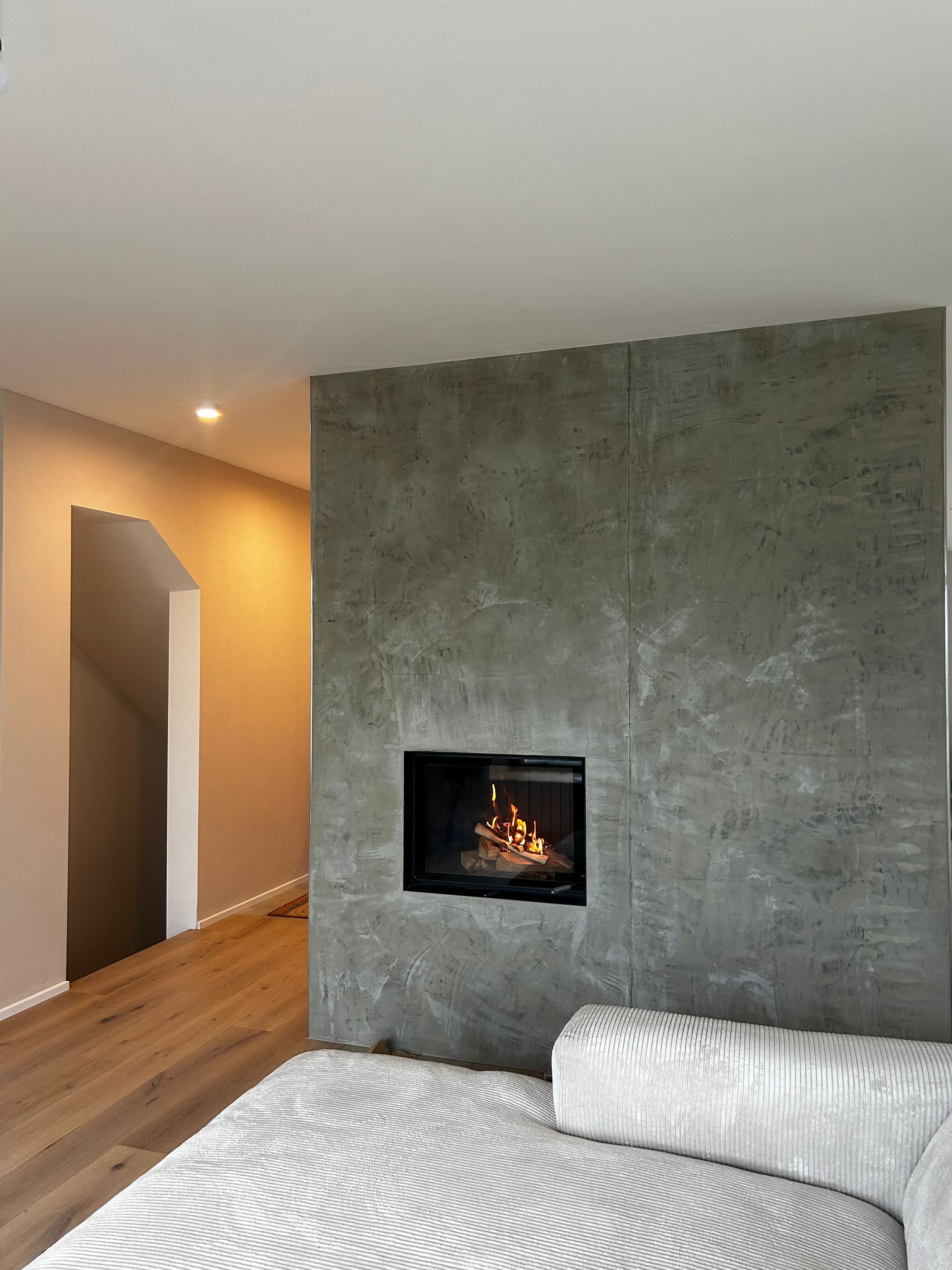 Ein moderner Kamin ist in eine strukturierte graue Wand eingelassen, neben einem Flur, über einem weißen Sofa.