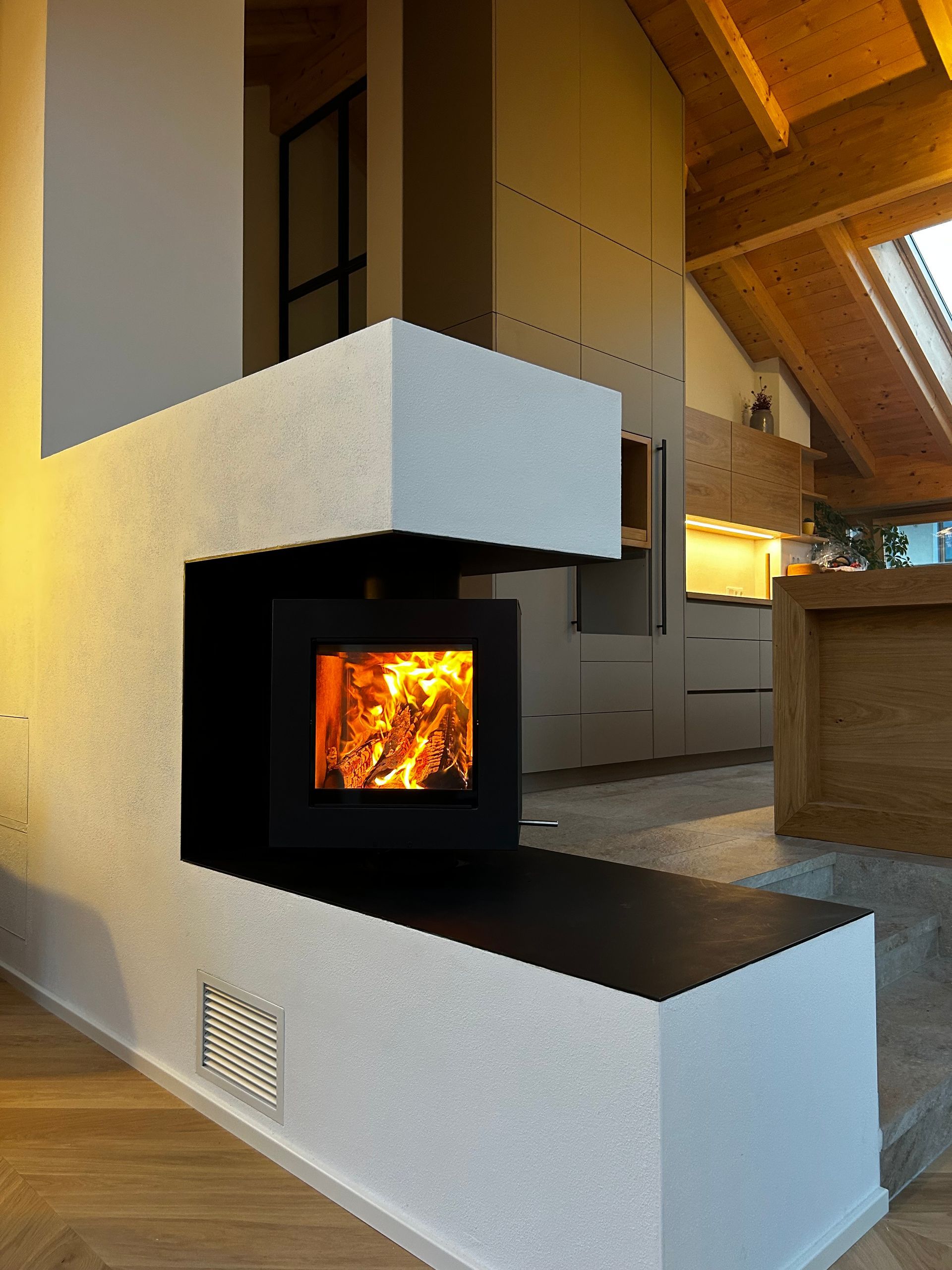 Moderner Kamin mit schwarz umrahmtem Feuerraum, eingebaut in eine weiße, schräge Steinkonstruktion.