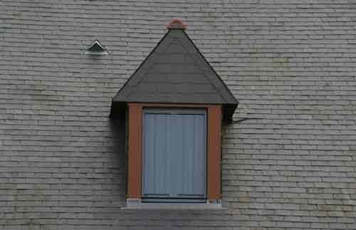 Lucarne avec toit pointu brun foncé et fenêtre à panneaux verticaux gris encastrée dans un toit en bardeaux gris.