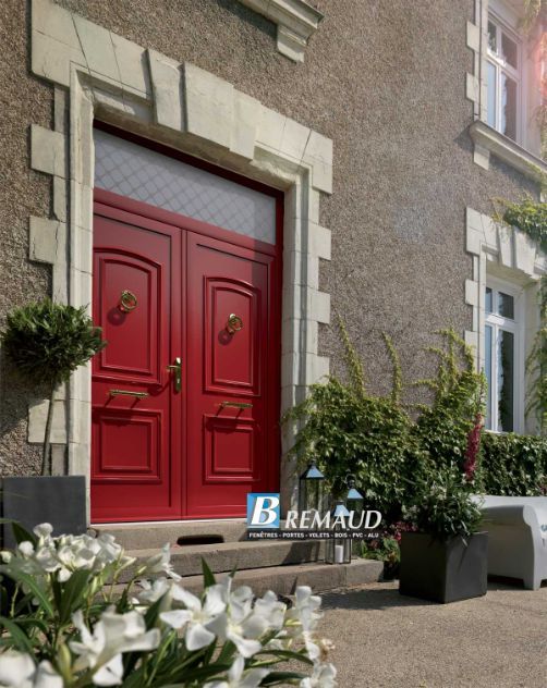 Double porte rouge avec encadrement en pierre sur une maison en pierre entourée de verdure.