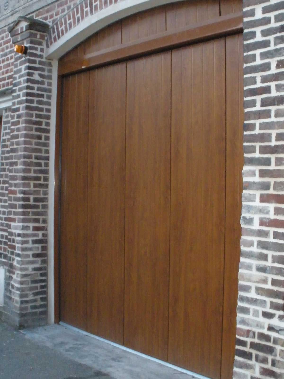 Porte de garage verticale en bois marron dans un bâtiment en briques.