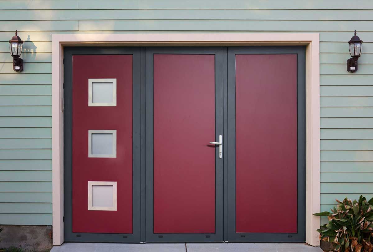 Portes de garage rouges et grises avec trois fenêtres carrées, encadrées de lumières sur un mur vert.