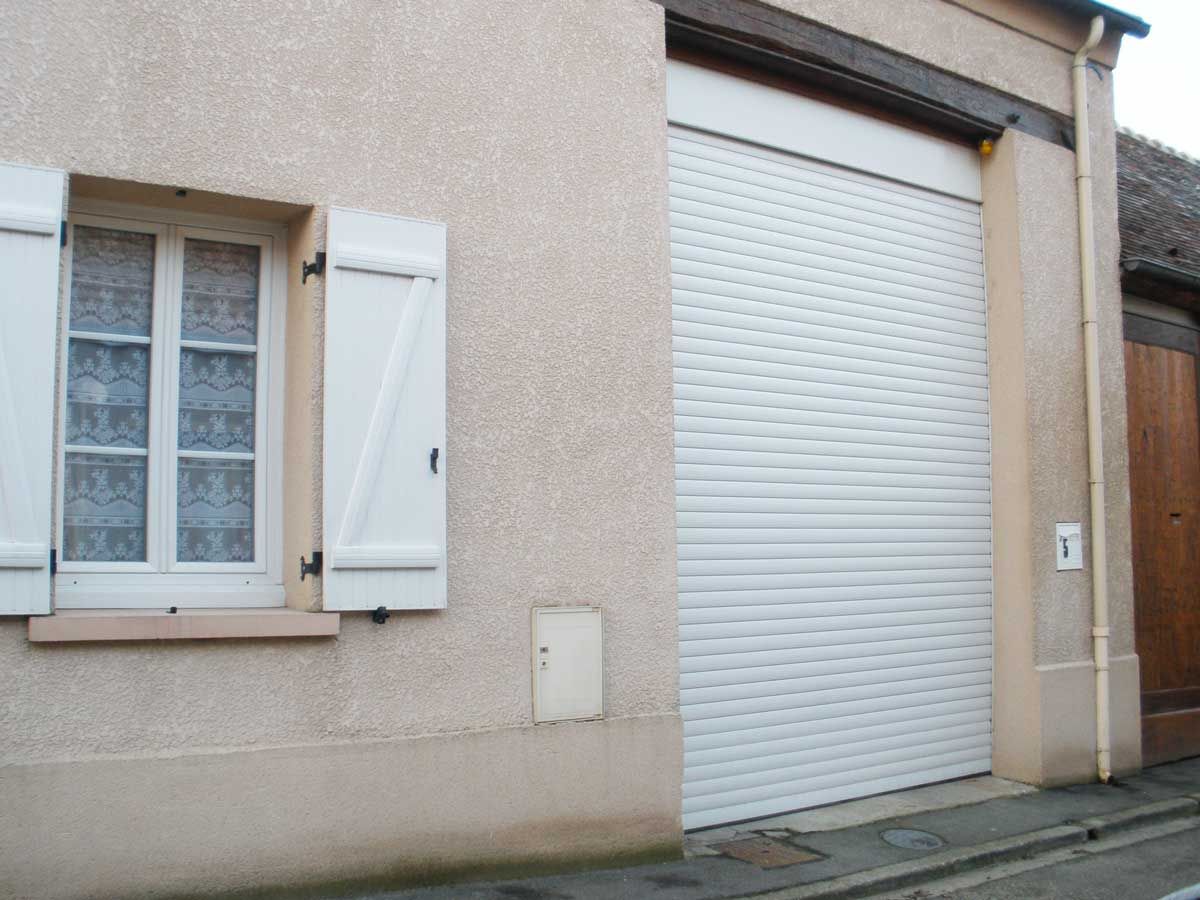 Bâtiment beige avec une fenêtre et des volets, et une porte sectionnelle blanche fermée.
