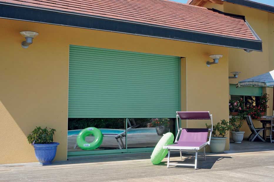 Un volet roulant turquoise recouvre partiellement une fenêtre donnant sur un espace piscine aménagé avec une chaise longue, un matelas gonflable et des plantes.