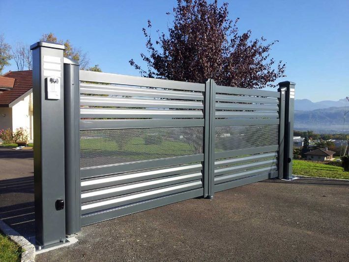 Portail d'allée en métal gris, à lattes horizontales et motif grillagé. Installé devant une maison, il donne sur un paysage verdoyant.
