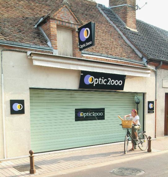 Magasin Optic 2000 avec un volet vert fermé, une personne à vélo et une signalétique noire, blanche et violette.