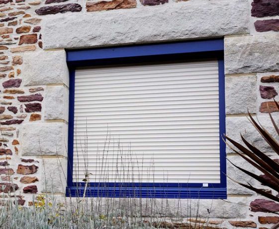 Fenêtre à volet roulant beige et cadre bleu foncé encastrée dans un mur en pierre.