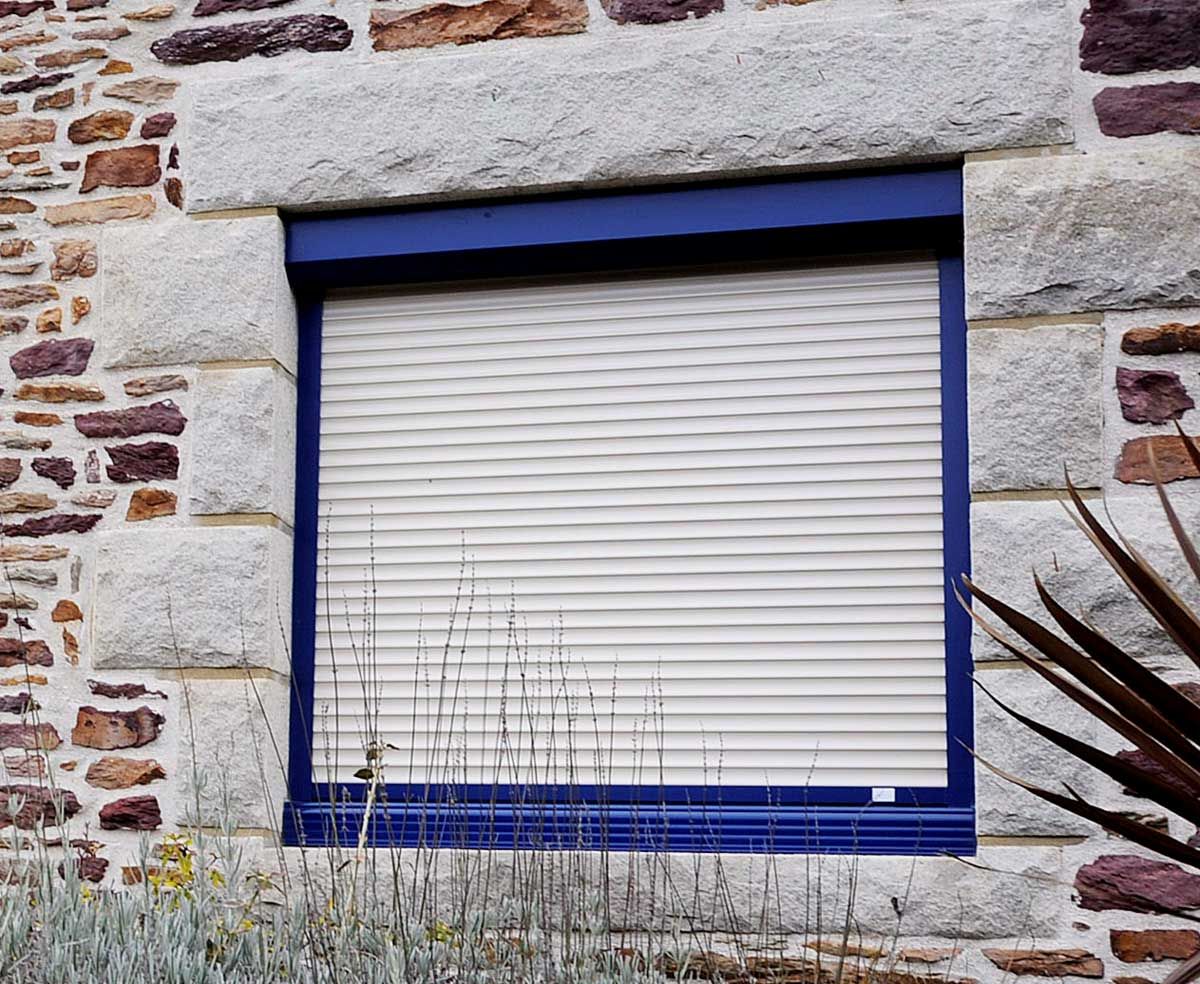 Fenêtre à cadre bleu avec volets roulants beiges fermés, dans un mur en pierre.