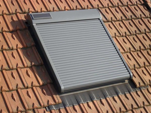 Volet roulant gris installé sur un toit en pente recouvert de tuiles brun-rouge.